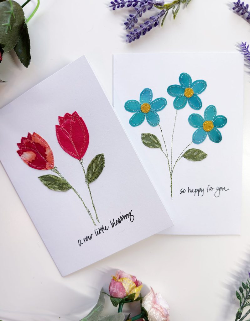 Two handmade greeting cards decorated with fabric flowers; one card has red tulips and the other has blue daisies. Handwritten messages read "a new little blessing" and "so happy for you.