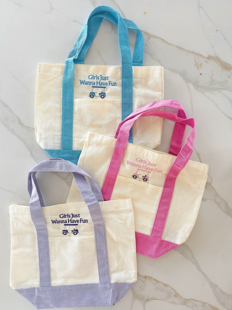 Three canvas tote bags with colored handles and trim (turquoise, pink, and lavender) are displayed. Each has the text "Girls Just Wanna Have Fun" and a small car graphic on the front pocket.