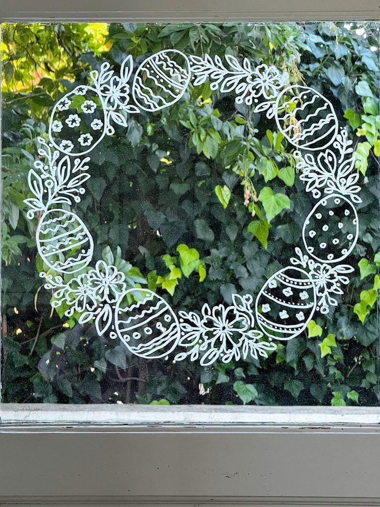 A white line drawing of a wreath made of decorated Easter eggs and leaves is displayed on a window, with green foliage visible outside.