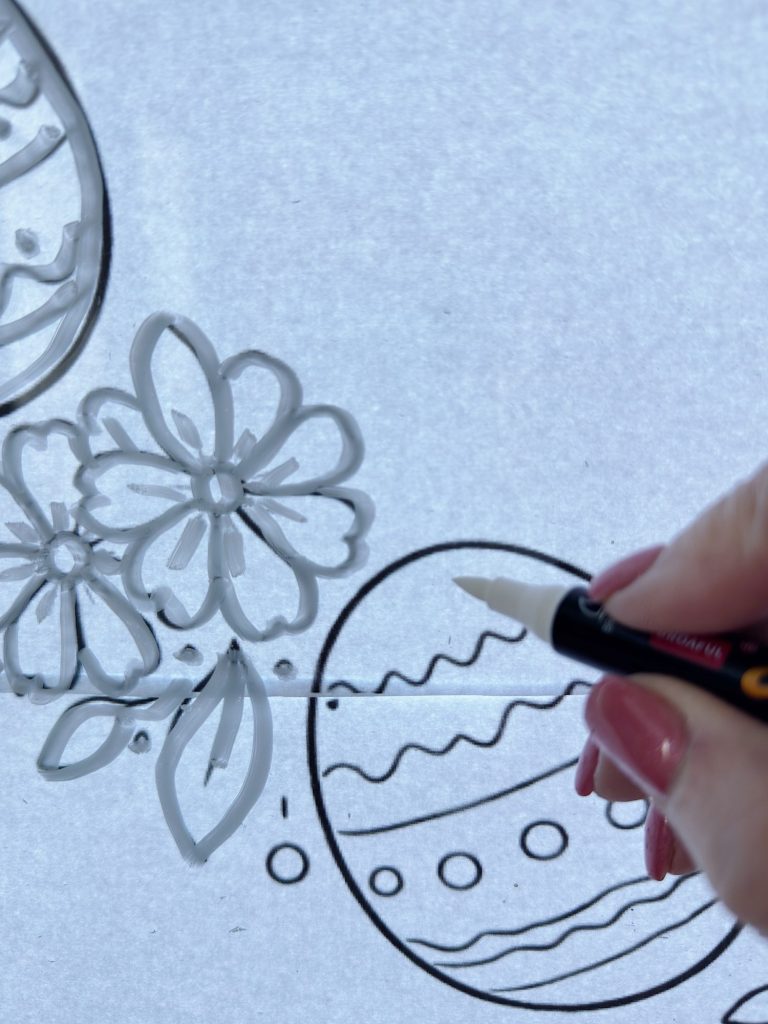 A hand with pink nails uses a black marker to outline flower and Easter egg designs on a white surface.