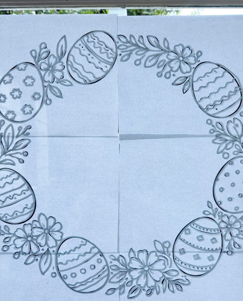 A circular wreath design made of decorated Easter eggs and flowers, drawn on four sheets of white paper taped together on a window.