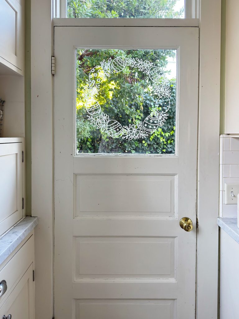 A white kitchen door with a glass window features a decorative white floral wreath design; greenery and sunlight are visible outside.