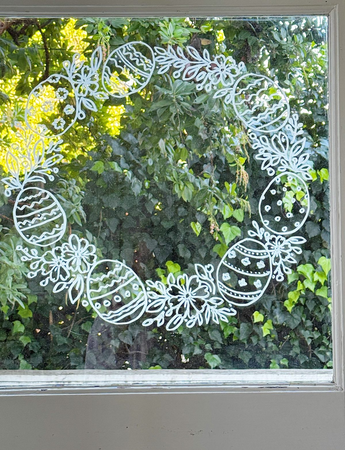 White line drawing of a wreath with flowers and decorated eggs on a window, with green foliage visible outside.