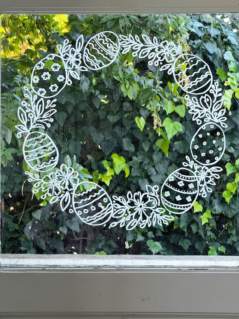 A circular wreath of hand-drawn Easter eggs and foliage is drawn with white marker on a window, with green leaves visible outside.