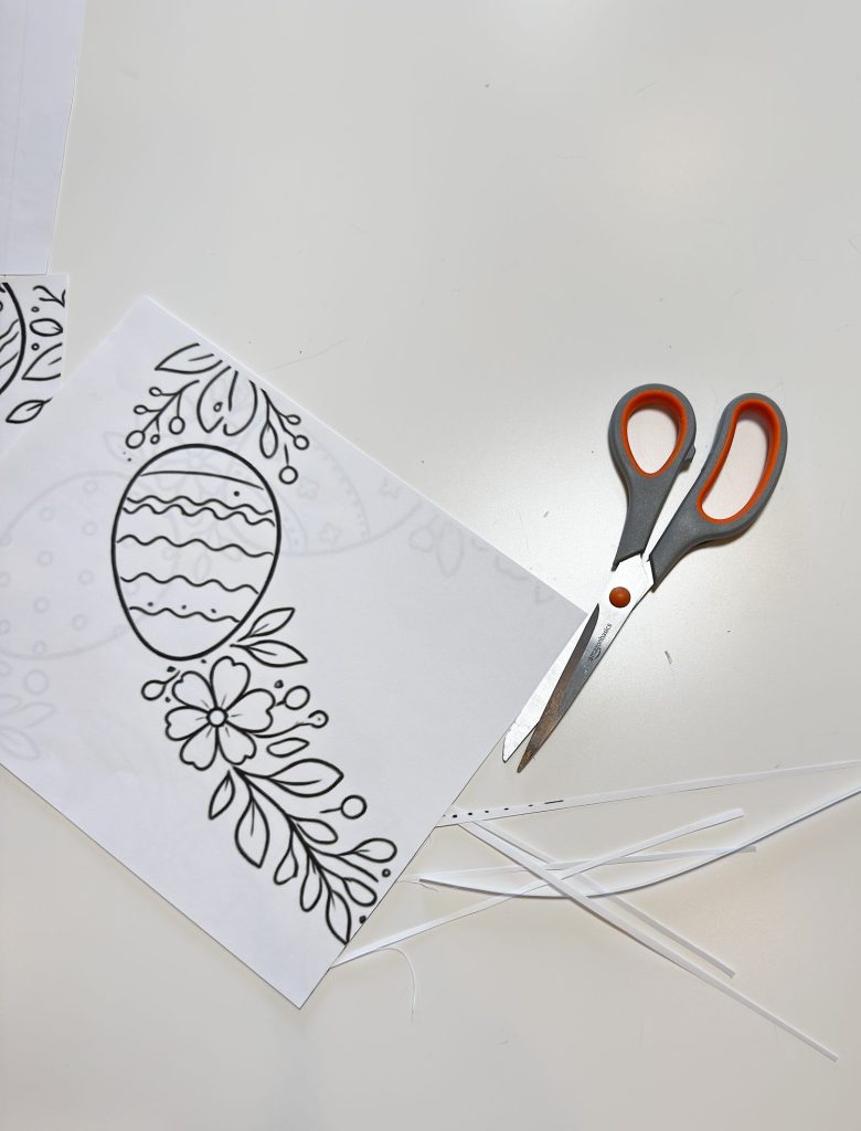 A pair of scissors and a sheet of paper with an Easter egg and floral design are on a white surface, with paper strips scattered nearby.