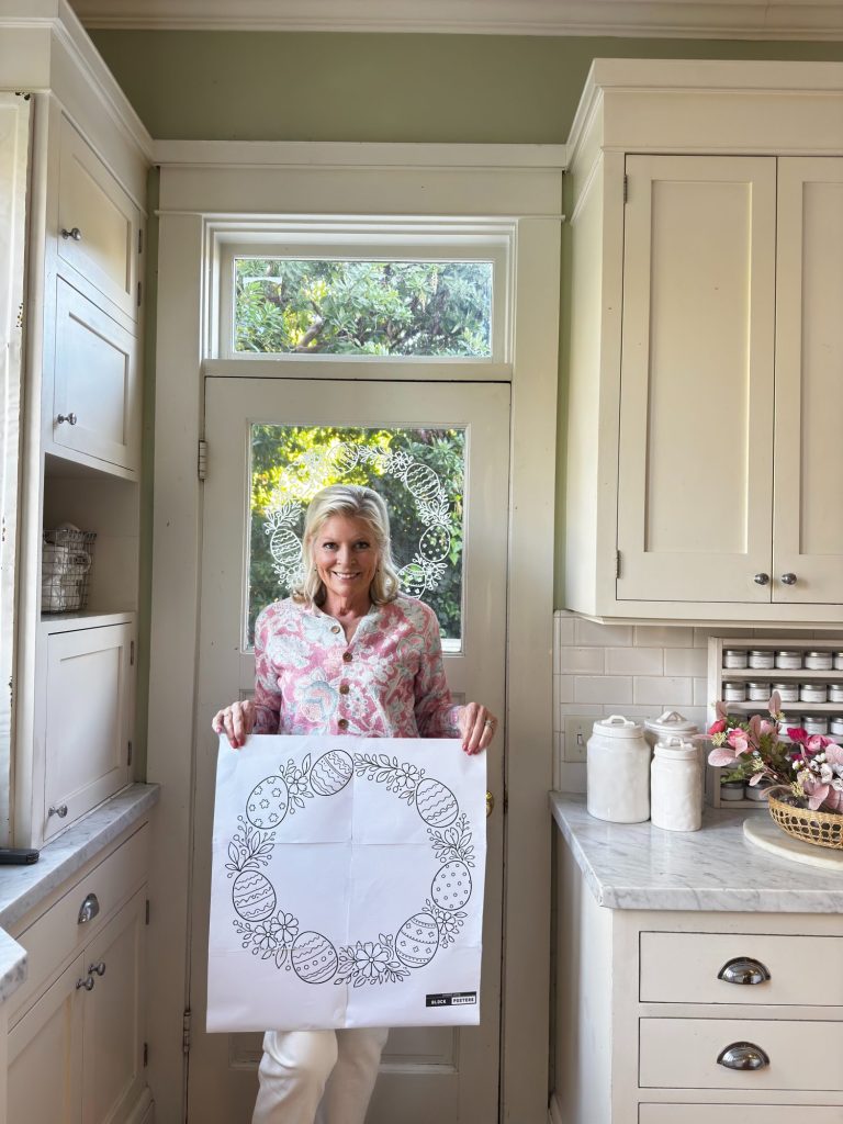 A woman stands in a kitchen, holding a large sheet of paper with a circular, decorative line drawing.