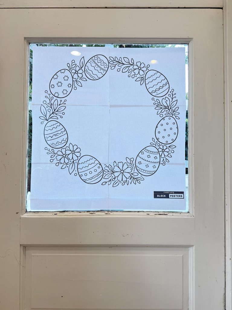 A line drawing of a wreath made of decorated Easter eggs and flowers is taped to a window on a white door.