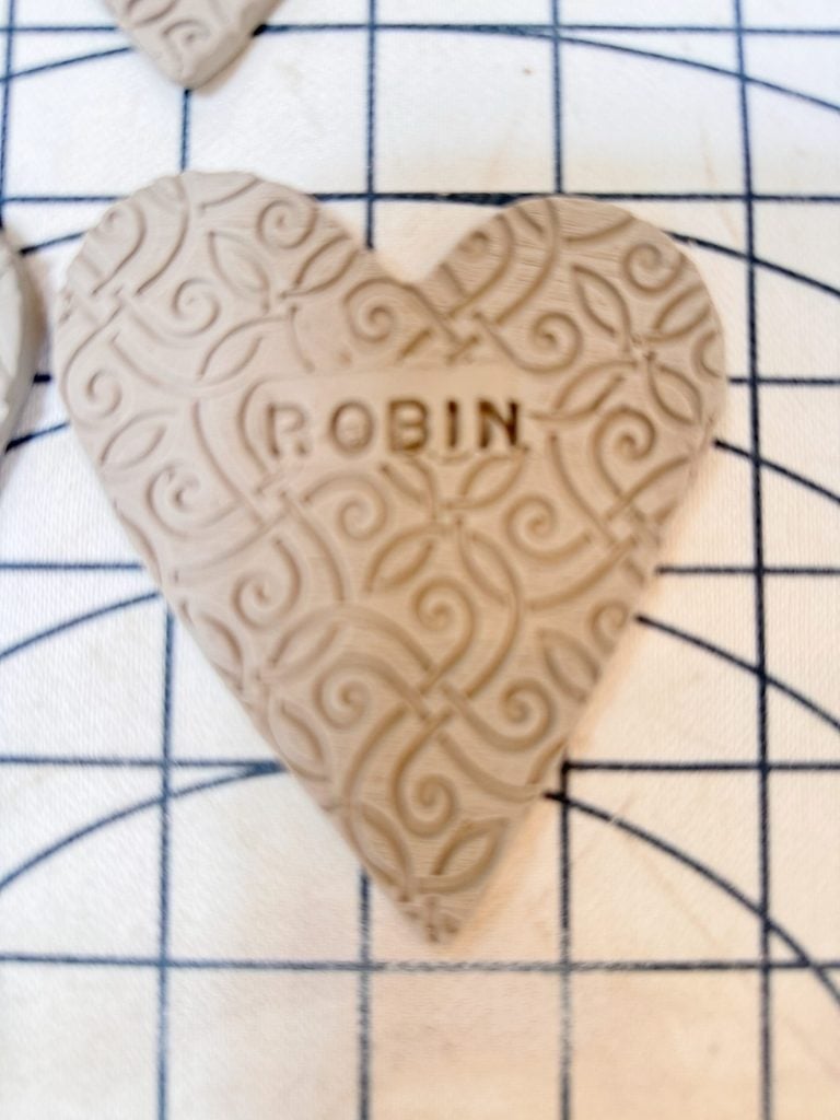 A heart-shaped piece of embossed clay with the name "ROBIN" stamped in the center, placed on a gridded fabric surface.