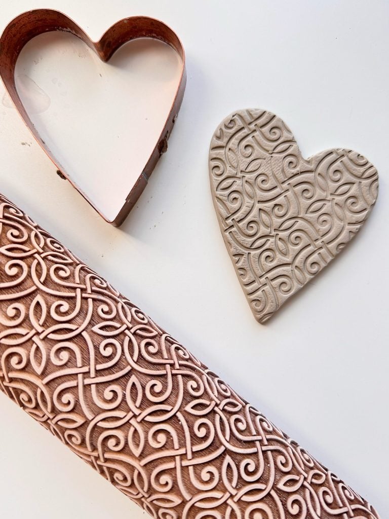 A heart-shaped clay piece with an intricate pattern, a metal heart-shaped cutter, and a patterned rolling pin on a white surface.