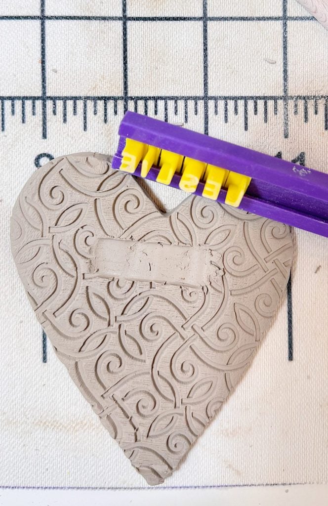 A textured clay heart with a rectangular indentation is beneath a purple tool with yellow letters on a grid mat.