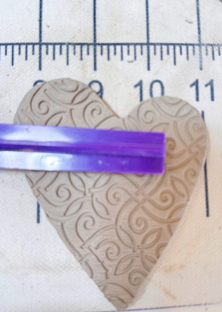 A textured clay heart beneath a purple tool is placed on a fabric surface marked with measurement lines.