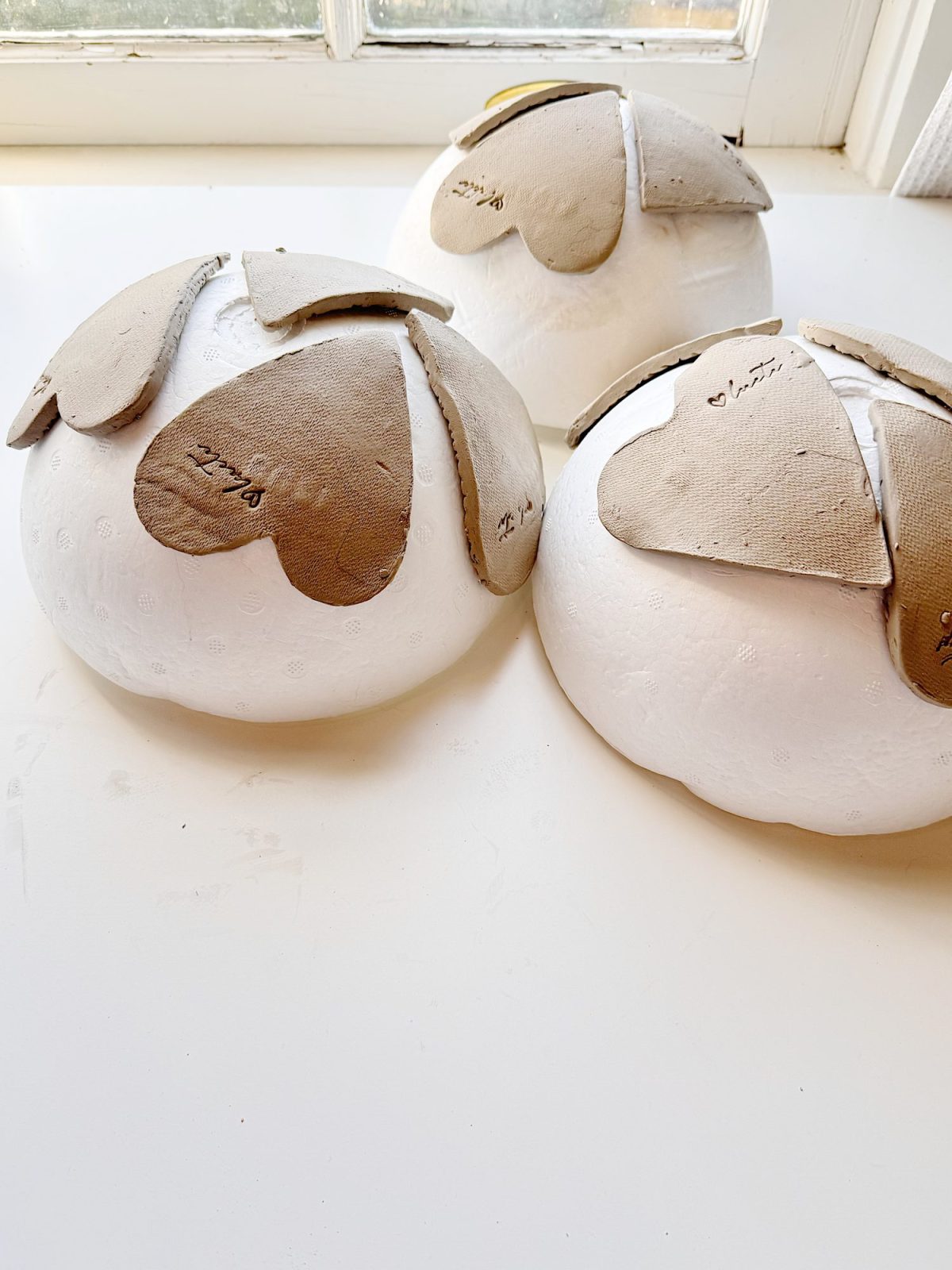 Three white spherical objects with textured surfaces, each partially covered by overlapping brown cardboard pieces, are placed on a white surface near a window.