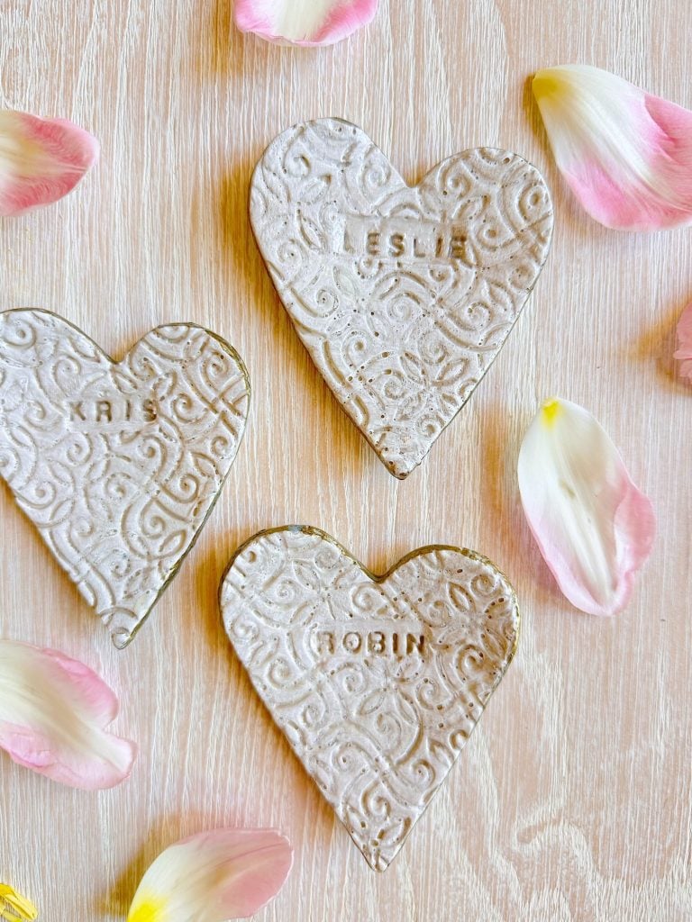 Three textured, heart-shaped clay ornaments with the names "KRIS," "LESLIE," and "ROBIN" stamped on them, surrounded by scattered rose petals on a light wooden surface.