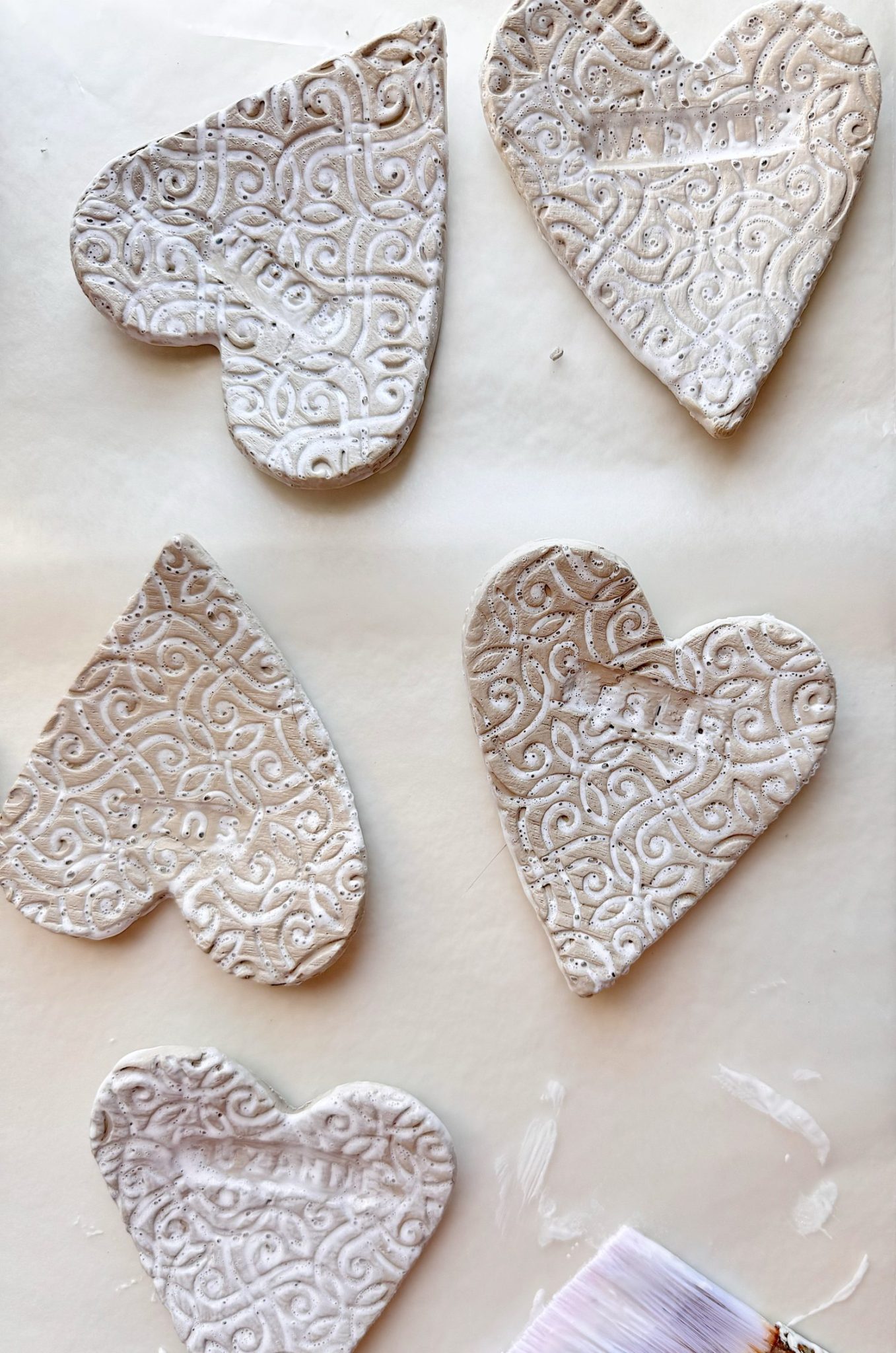 Five heart-shaped clay ornaments with embossed patterns are laid out on a white surface, with a paintbrush and some white paint visible at the bottom.
