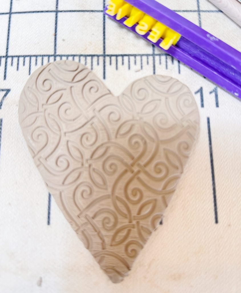 A clay heart with an embossed swirling pattern rests on a ruler-marked fabric surface, with purple and yellow plastic tools nearby.