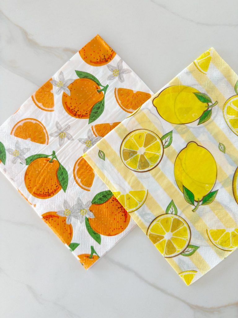 Two square paper napkins with fruit designs; one features oranges and white flowers, the other shows lemons and lemon slices with a yellow-striped background.