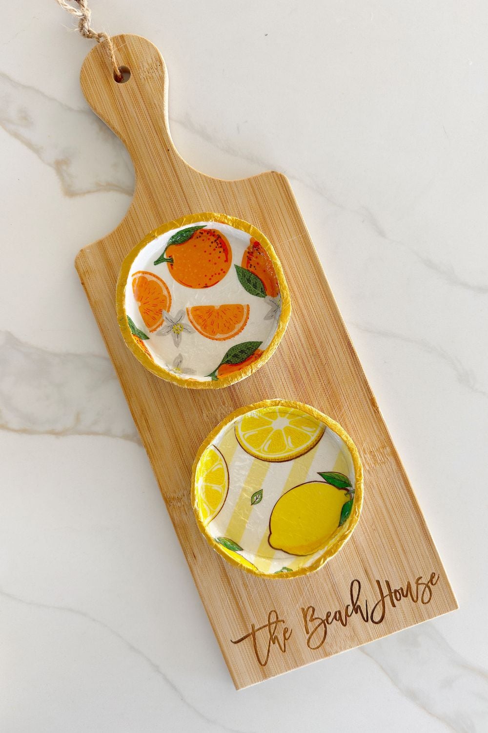 A wooden board with &ldquo;The Beach House&rdquo; engraved, holding two small dishes&mdash;one with an orange pattern and one with a lemon pattern&mdash;on a marble surface, beside a set of charming handmade cards.