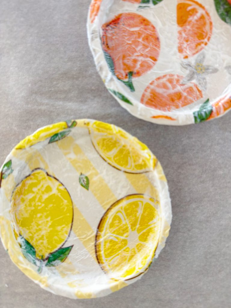 Two round bowls covered with plastic wrap, one with a lemon design and the other with an orange design, placed on a gray surface.