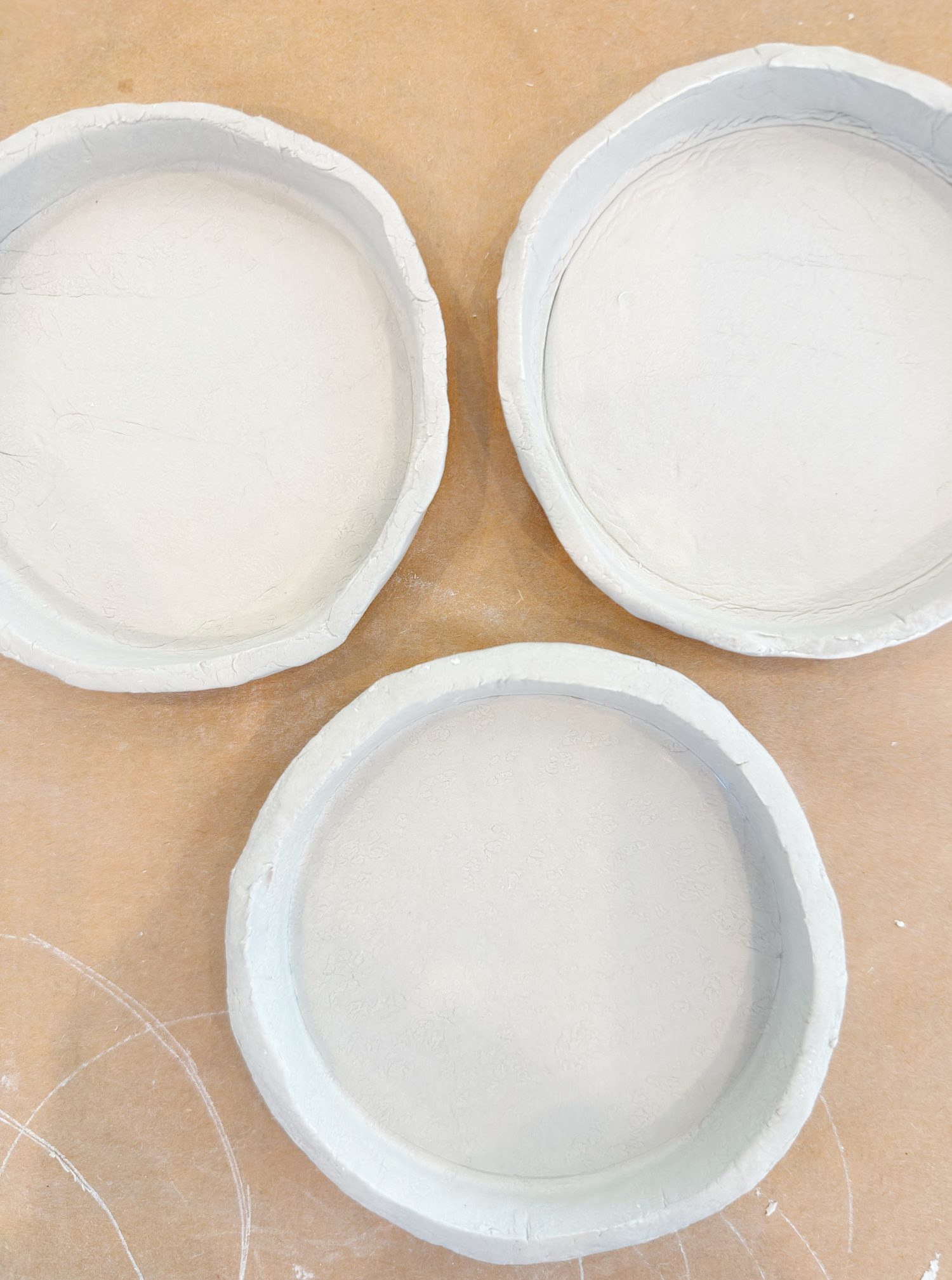 Three round, shallow, unfinished clay dishes are arranged on a light brown surface.