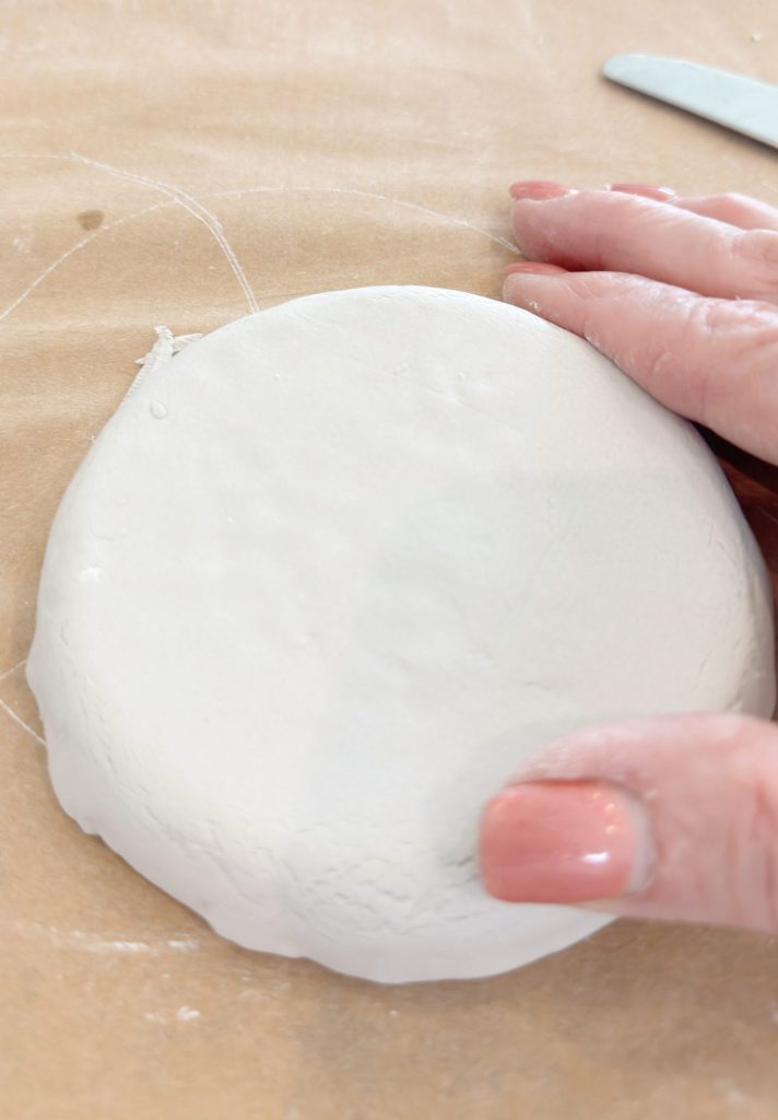 A hand pressing down on a round, flat piece of white clay resting on brown parchment paper.