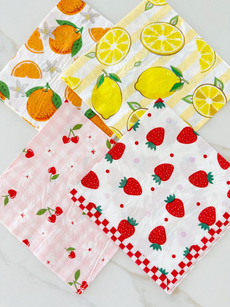 Four square napkins with fruit patterns: oranges, lemons, cherries, and strawberries, arranged in a grid on a white surface.
