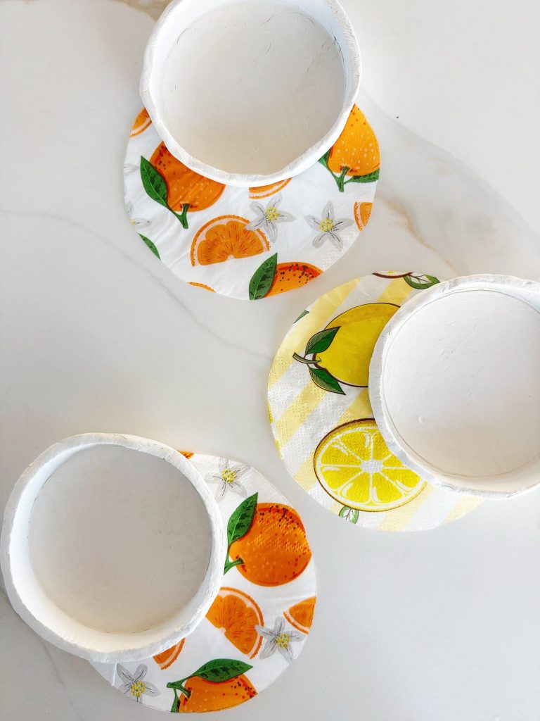 Three white bowls are placed upside down on a white surface, each resting on a colorful, fruit-themed napkin with orange and lemon designs.