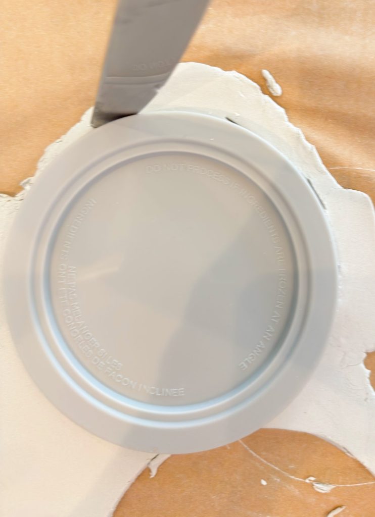 A round plastic lid placed upside down on a sheet of white clay, with a knife cutting along the edge of the lid.