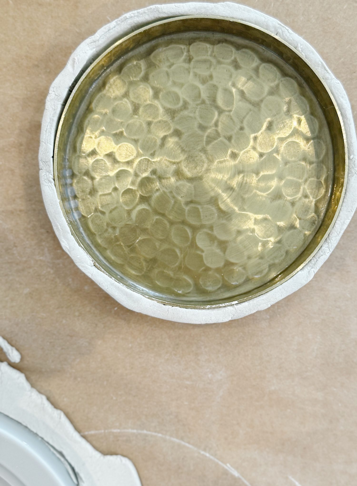 A round, hammered brass dish set inside a white, uneven plaster base on a light brown surface.