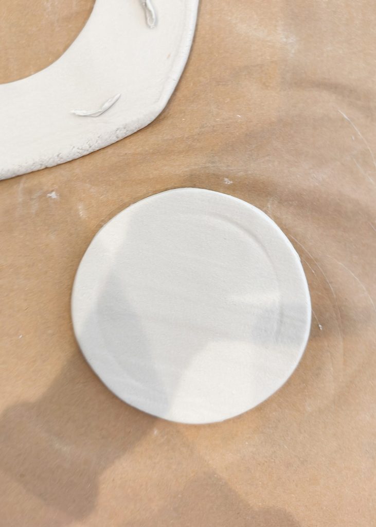 A round, flat piece of white clay sits on a brown surface, with a partial view of another clay piece nearby.