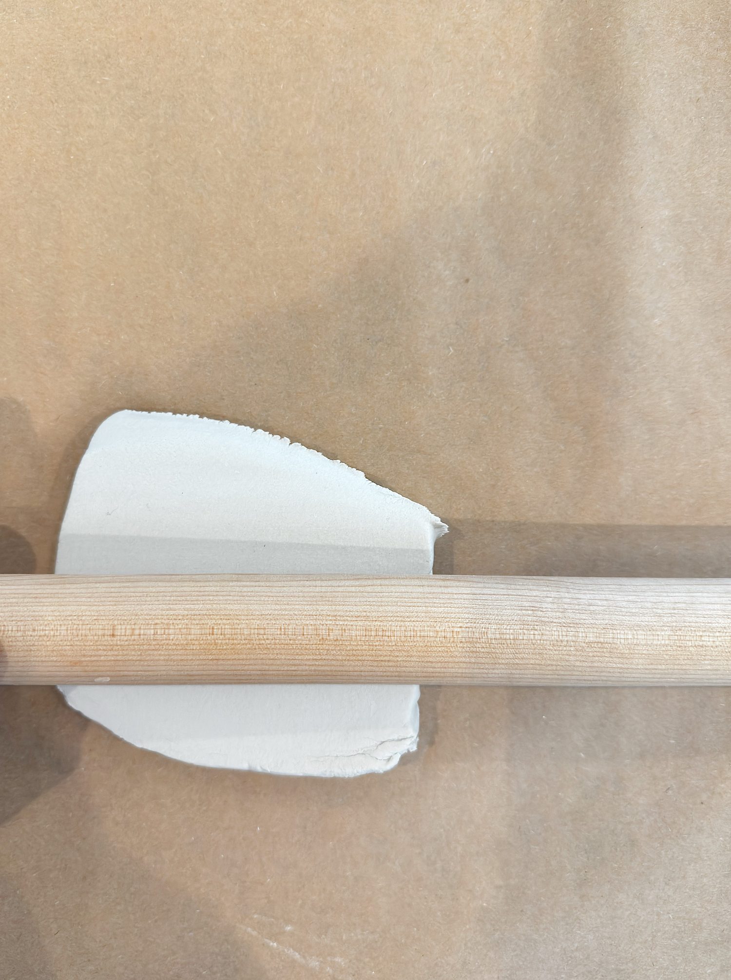 A wooden rolling pin flattening a piece of white clay on brown parchment paper.
