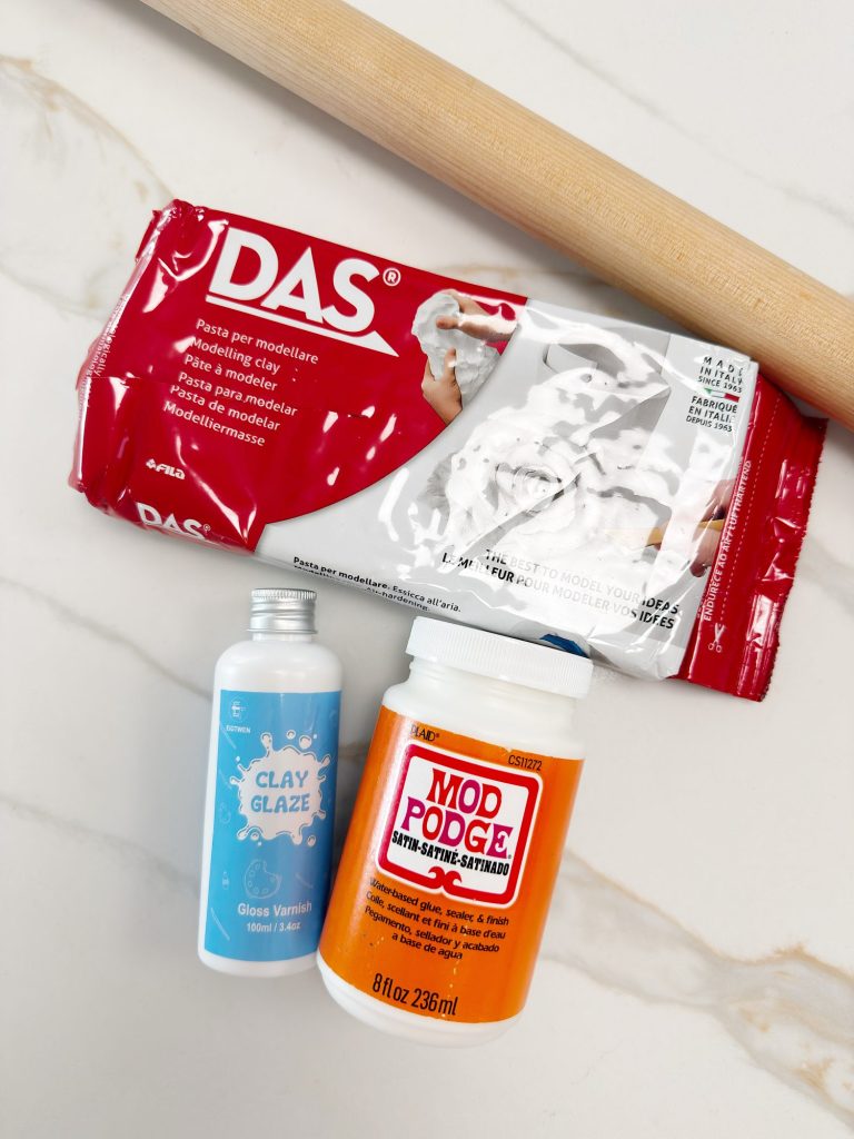 A pack of DAS modeling clay, a bottle of clay glaze, a container of Mod Podge, and a wooden rolling pin on a white surface.