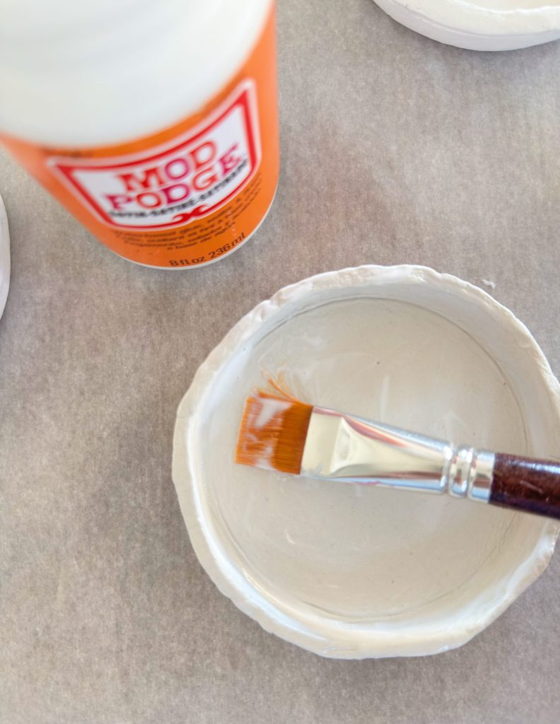 A paintbrush rests in a small, round ceramic dish while a bottle of Mod Podge sits nearby on a gray surface.