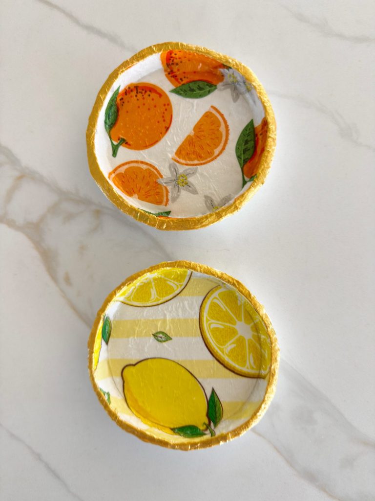 Two small, round dishes with gold rims: one decorated with oranges and leaves, the other with lemons and yellow stripes, placed on a white marble surface.