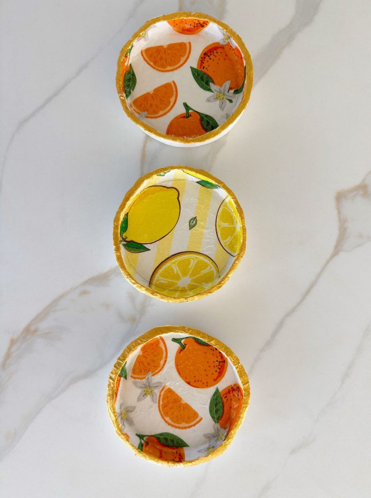 Three small round bowls with fruit-themed designs—orange slices on the top and bottom bowls, and lemons on the middle bowl—are placed on a white marble surface.