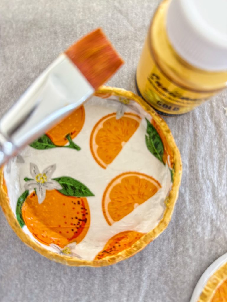 A small dish with an orange slice pattern, a paintbrush, and a bottle of gold paint are placed on a grey surface.