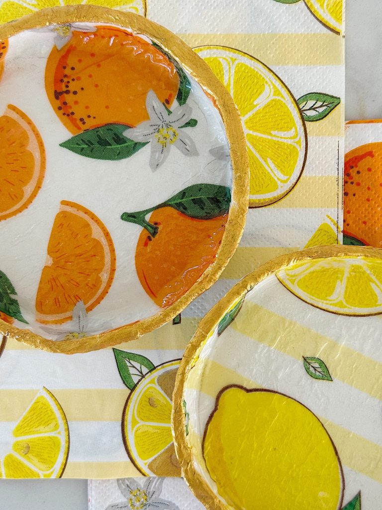 Round dishes with gold rims featuring painted oranges, lemons, and flowers, placed on napkins decorated with similar citrus fruit illustrations.