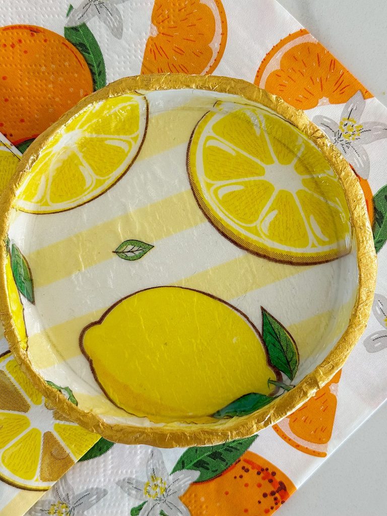 A round paper plate with a yellow lemon and lemon slices design, featuring a gold rim, placed on orange and lemon-themed napkins.