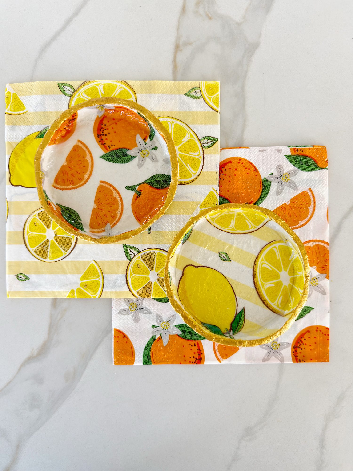 Two cocktails with citrus fruit garnishes on citrus-themed napkins featuring orange and lemon designs, placed on a white marble surface.