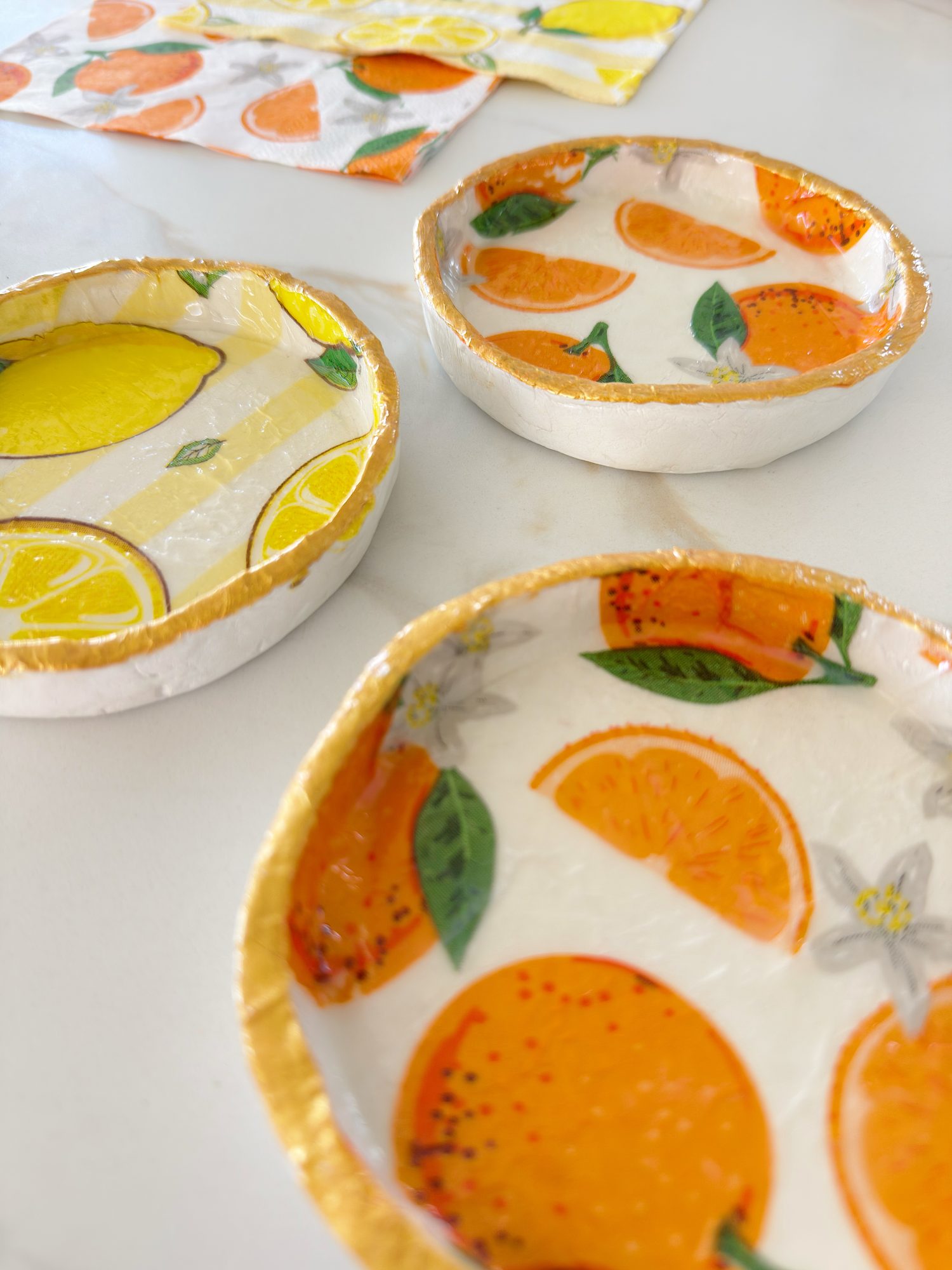 Three small round dishes with fruit designs&mdash;one with lemons, two with oranges&mdash;are displayed on a white surface. A fruit-patterned napkin is visible in the background.