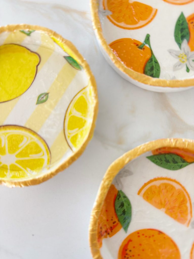 Three small round dishes with colorful citrus fruit designs, including lemons and oranges, displayed on a white marble surface.