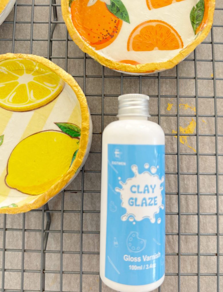 A bottle of clay glaze sits on a wire rack next to two circular clay dishes decorated with painted citrus fruit designs.