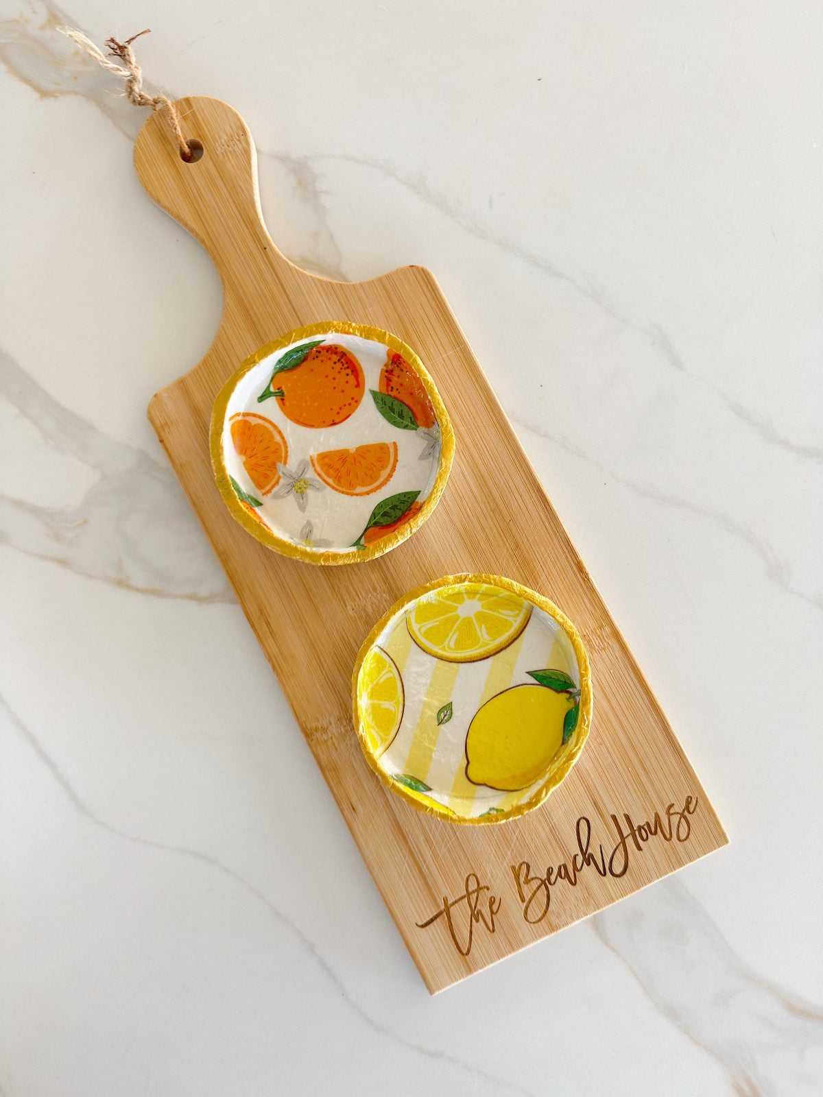 A wooden serving board with "The Beach House" engraved, holding two small round plates with citrus fruit designs, one with oranges and one with lemons.