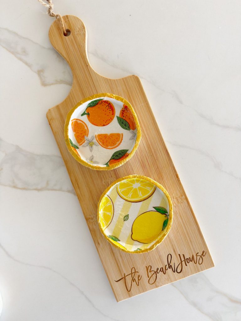 A wooden cutting board labeled "The Beach House" holds two round resin coasters with orange and lemon fruit designs, placed on a white marble surface.