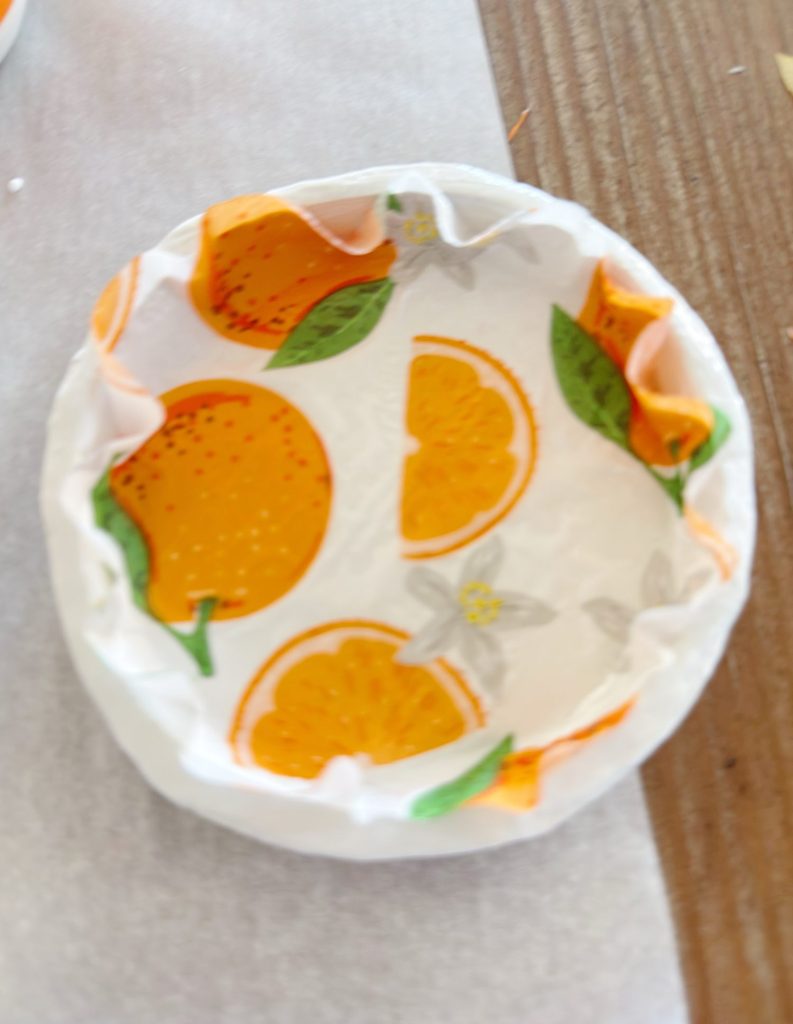 A round dish lined with parchment paper printed with orange fruit and green leaf illustrations sits on a wooden surface.