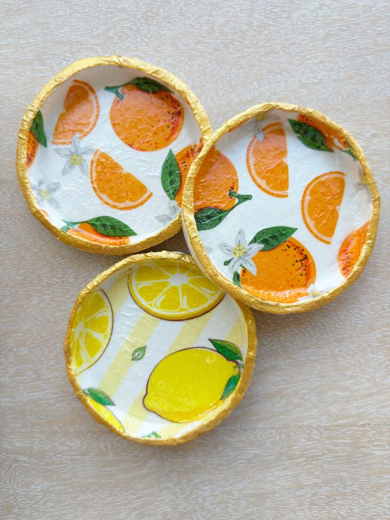 Three small round dishes with gold rims, each featuring a different citrus fruit design: oranges, lemons, and orange slices, placed on a light-colored surface.