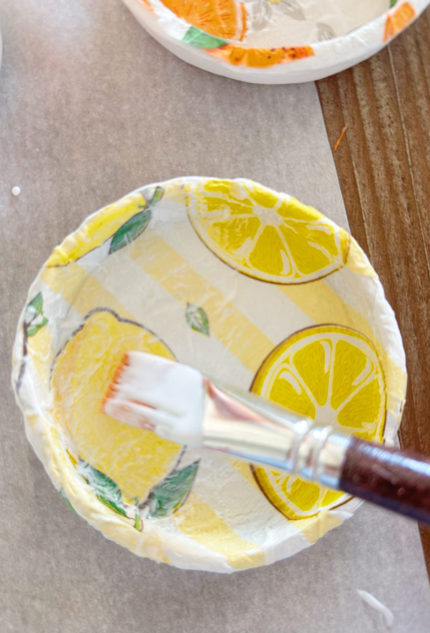 A paintbrush applies glue or sealant to a small paper bowl decorated with yellow lemon slice designs.