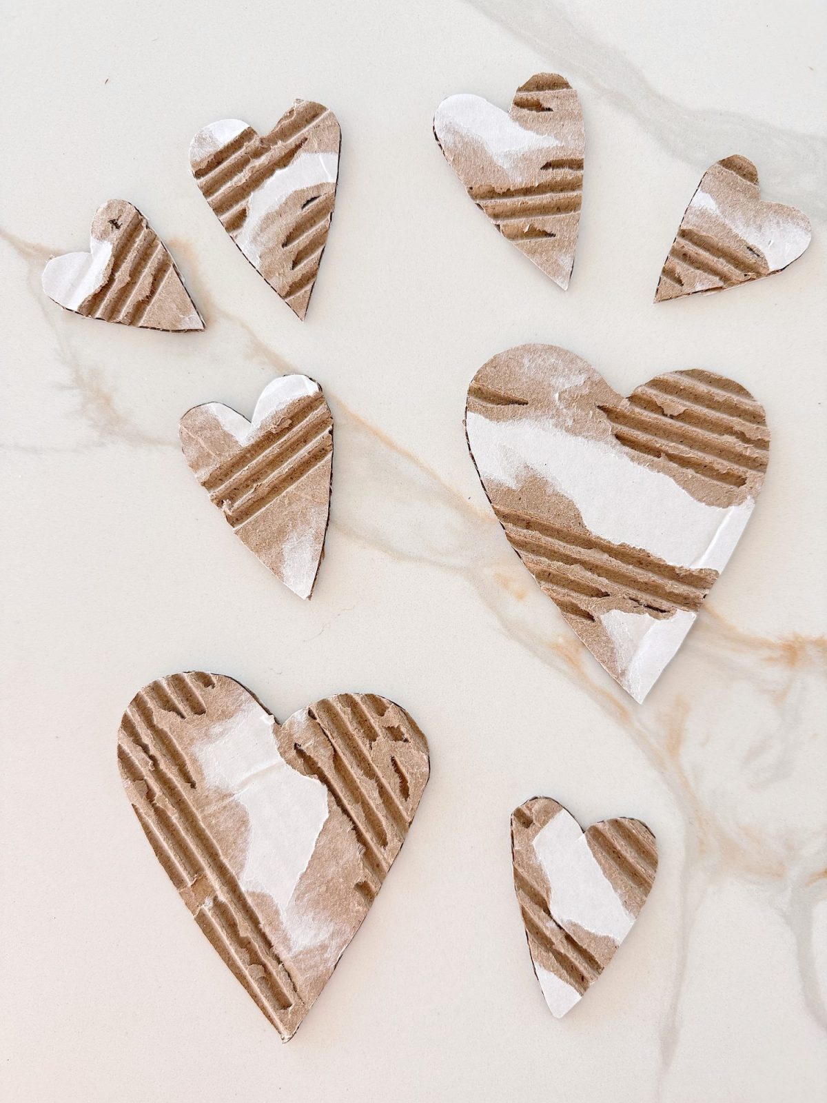 Eight heart-shaped cutouts made from corrugated cardboard with white paint accents are arranged on a light-colored surface.