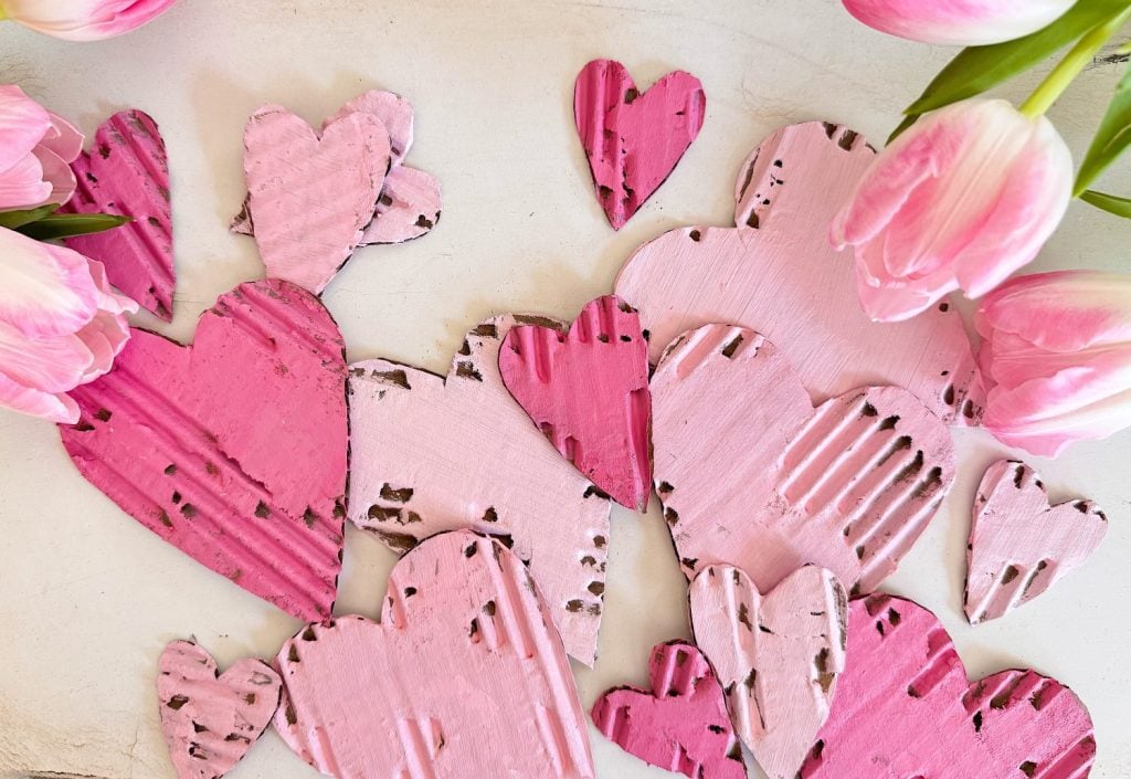 Several pink and light pink cardboard hearts arranged on a white surface, surrounded by pink tulips at the edges.