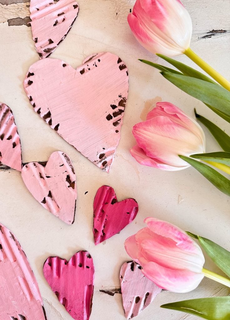 Pink and red painted wooden hearts scattered on a white surface next to three pink and white tulips with green stems.