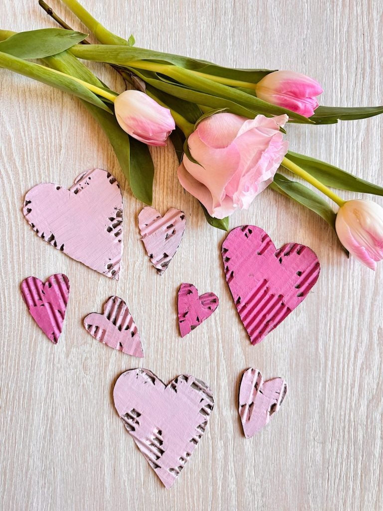 Pink tulips and a pink flower lie on a light wooden surface next to several pink, heart-shaped paper cutouts with various patterns.
